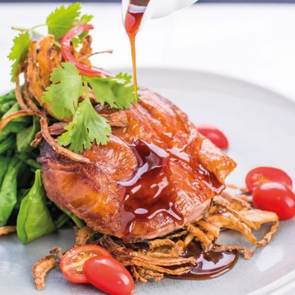 Crispy duck breast with sauce being poured, served with greens, fried onions, and cherry tomatoes.